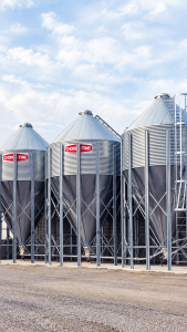 Three Chore-Time galvanized steel feed bins with conical roofs, hopper bottoms and support legs for bulk poultry feed storage.