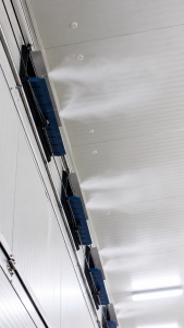 High-pressure misting nozzles releasing fine water mist along the ceiling of a livestock barn for evaporative cooling.